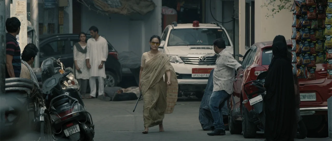 Nishaanchi 2 (2025) movie still: fullbody — A paved area behind buildings features several parked cars and a group of people. A woman in a tan sari…