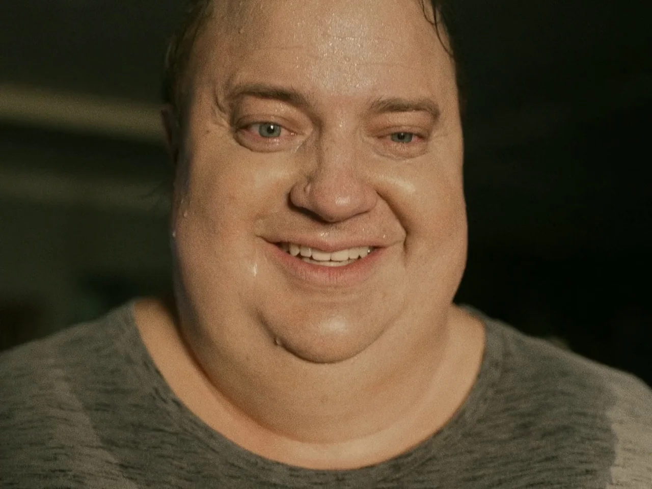 The Whale (2022) movie still: closeup — A man's face fills the frame in a dimly lit, dark indoor environment. The subject smiles at the viewer with…