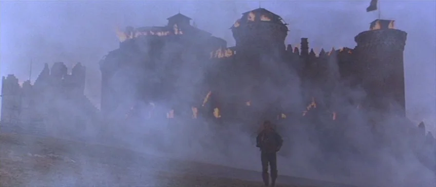 Flesh+Blood (1985) movie still: wide — A stone fortification burns with large flames while thick gray smoke covers the ground. A single man walks…