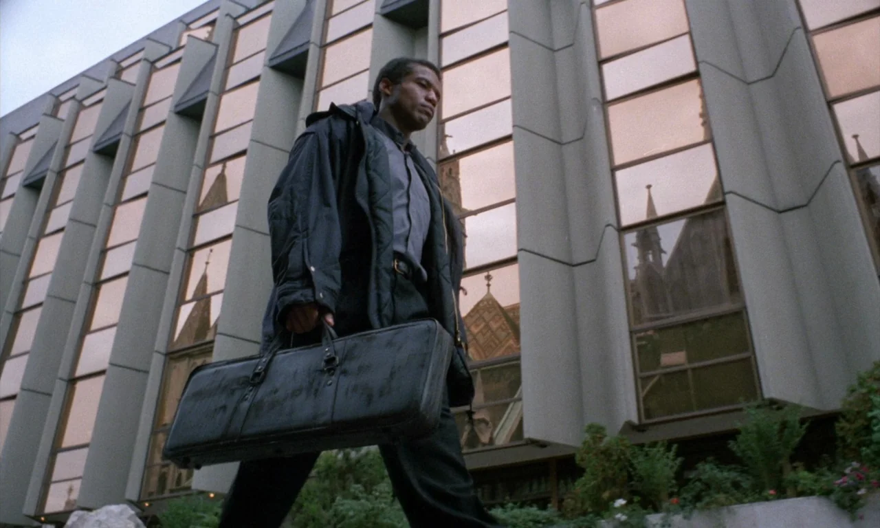 The Church (1989) movie still: fullbody — A modern building with reflective tinted glass windows stands behind a man walking past. The man carries a…