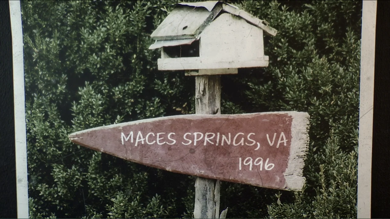 The Winding Stream (2014) movie still: closeup — A weather-beaten wooden signpost with text sits in front of thick, dark green shrubbery. The sign…