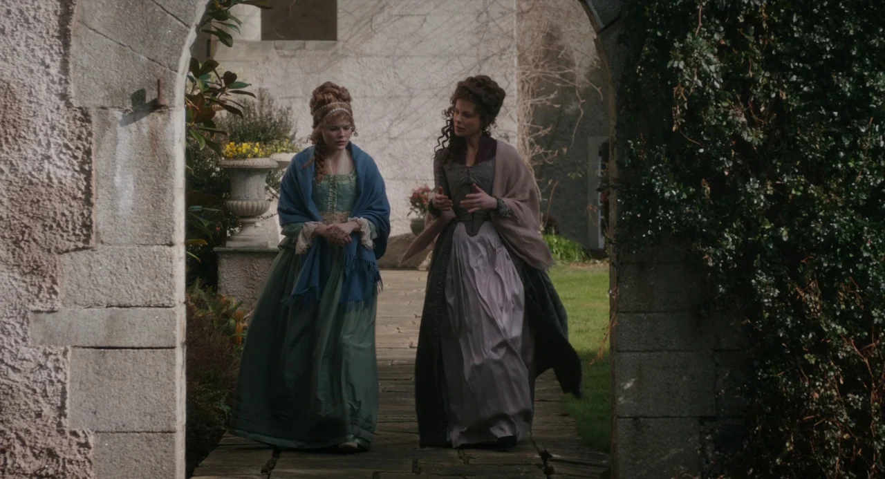 Love and Friendship (2016) movie still: fullbody — Two women in historical attire walk toward the camera through a stone archway. They move along a…