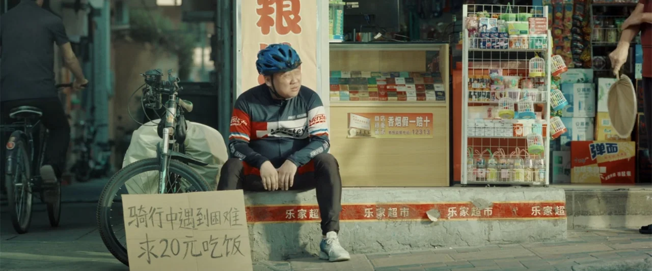 Yolo (2024) movie still: medium — A man sits on a concrete step outside a small retail shop with goods. He rests next to his bicycle and a…