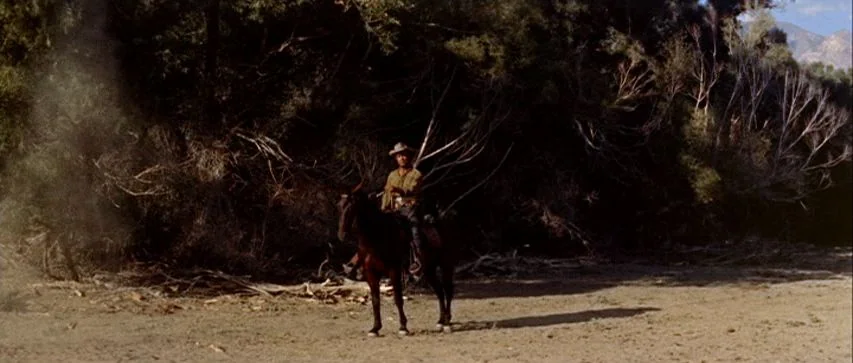 Ride Lonesome (1959) movie still: wide — A man on horseback stands before a dense thicket of bare and leafy trees. The landscape is a flat, dry,…