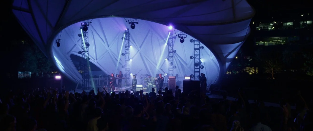 The Circle (2017) movie still: wide — An outdoor amphitheater features a modern white tensile structure illuminated by blue stage lights. A crowd of…