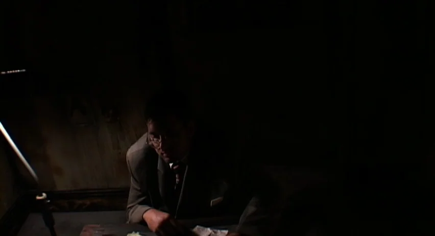 Inland Empire (2006) movie still: medium — An dimly lit, sparse room with a textured wall behind the subject. A man sits at a desk while illuminated…