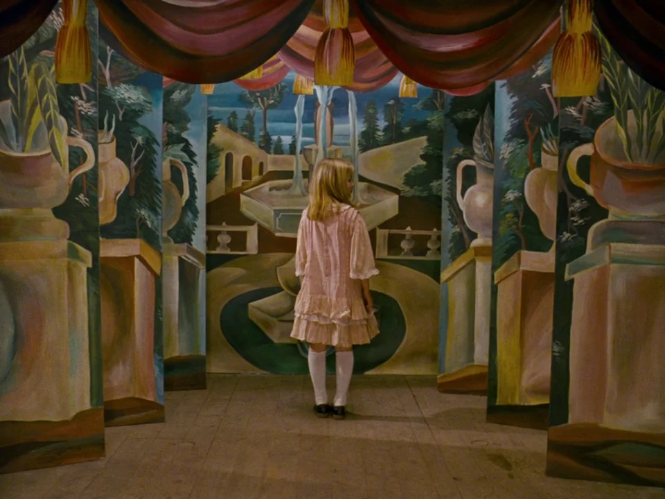 Alice (1988) movie still: fullbody — A girl stands centered in a small enclosure formed by hand-painted landscape panels. The backdrop features a…