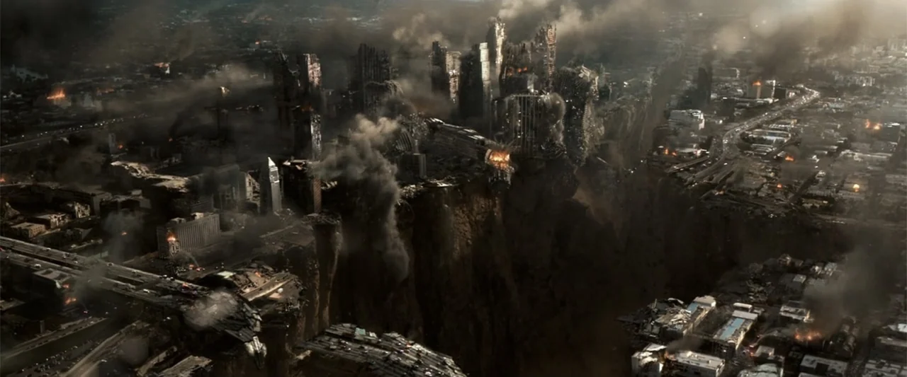2012 (2009) movie still: establishing — A decimated urban landscape split by a massive chasm showing collapsing high-rise buildings. Thick black…