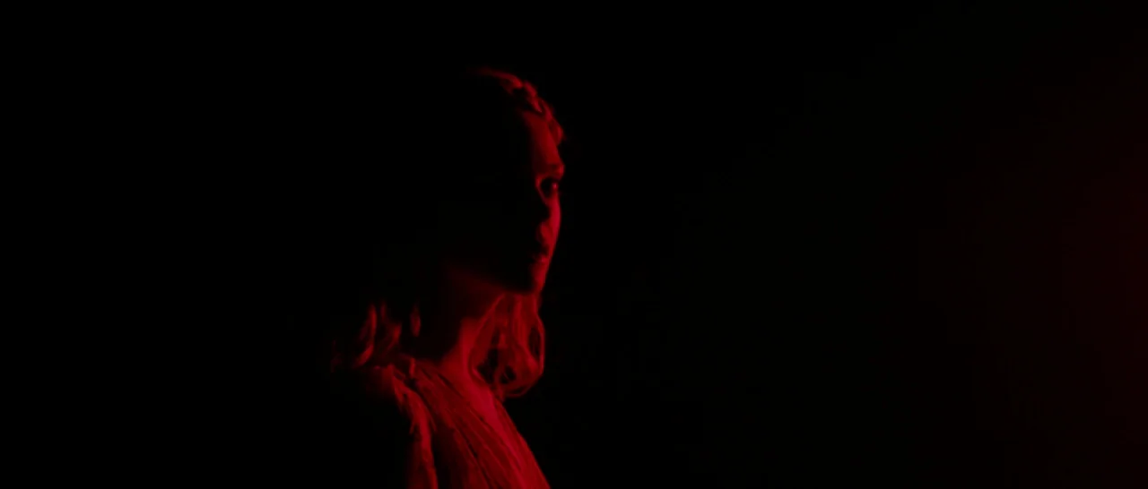 The Neon Demon (2016) movie still: medium — A figure stands in a dark space lit only by a deep red light. The woman stands alone, looking toward the…