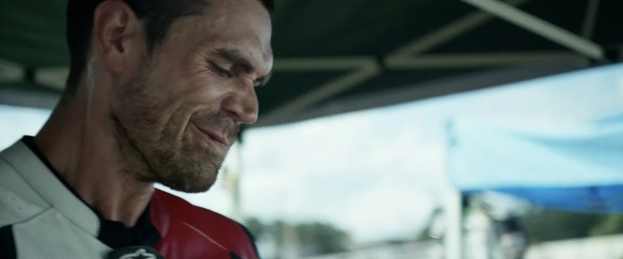 One Fast Move (2024) movie still: closeup — A sunny outdoor trackside area features a racing canopy in the blurred background. A man in a red and…