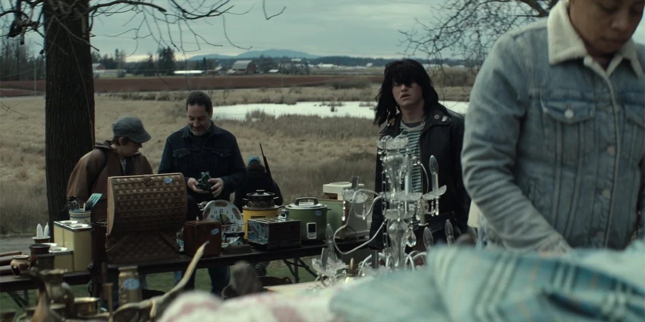 The Monkey (2025) movie still: medium — An outdoor flea market is set up in a field under an overcast sky. Several individuals browse through tables…