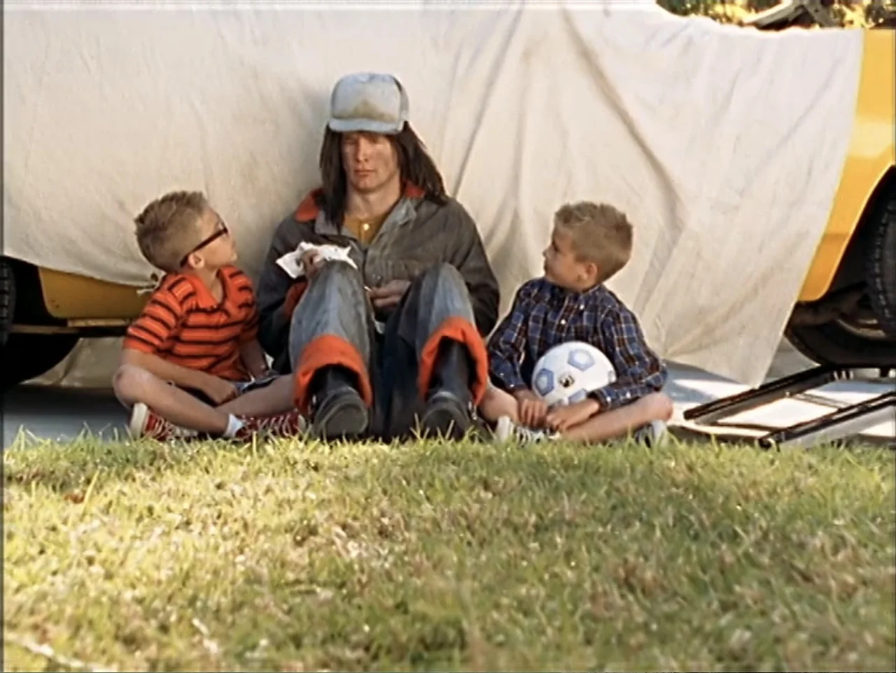 Just 4 Kicks (2003) movie still: fullbody — Three people sit on grass in front of a car covered by a white sheet. The two children look at the man…