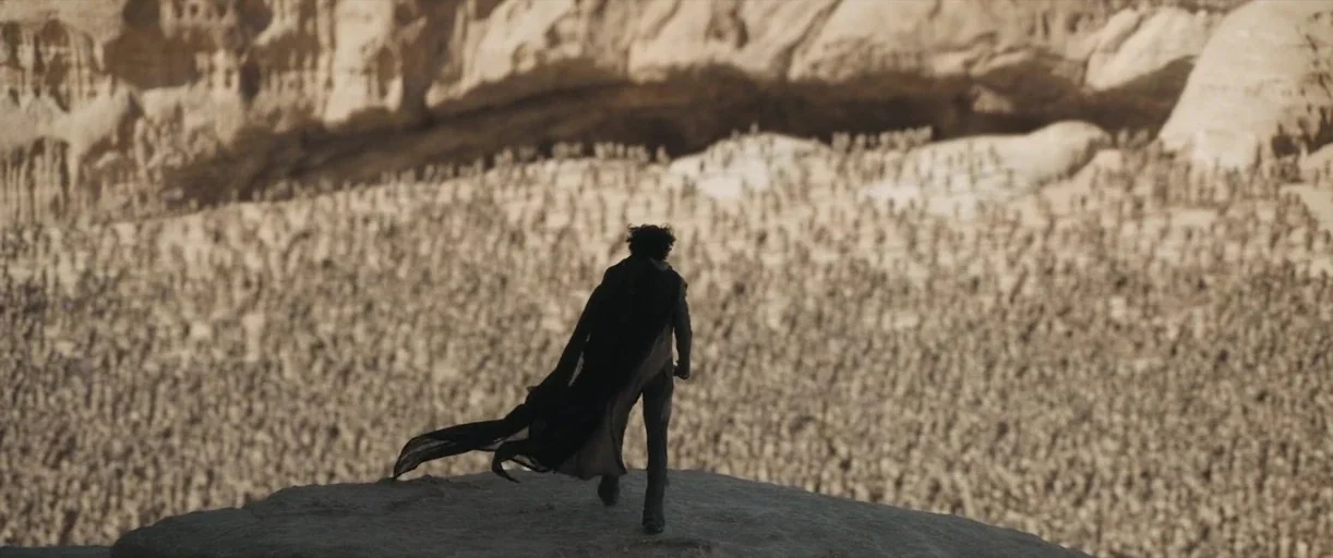 Dune: Part 2 (2024) movie still: wide — A desolate rocky desert landscape features a vast gathering of thousands of tiny figures. A lone man stands…
