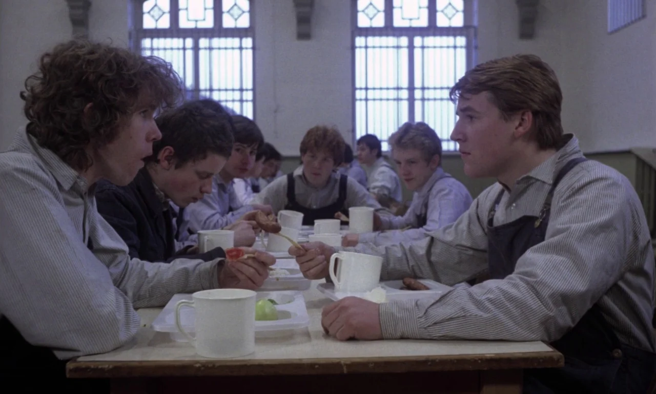 Scum (1979) movie still: medium — A utilitarian dining hall with tall barred windows and rows of long tables. Boys in striped shirts and dark aprons…