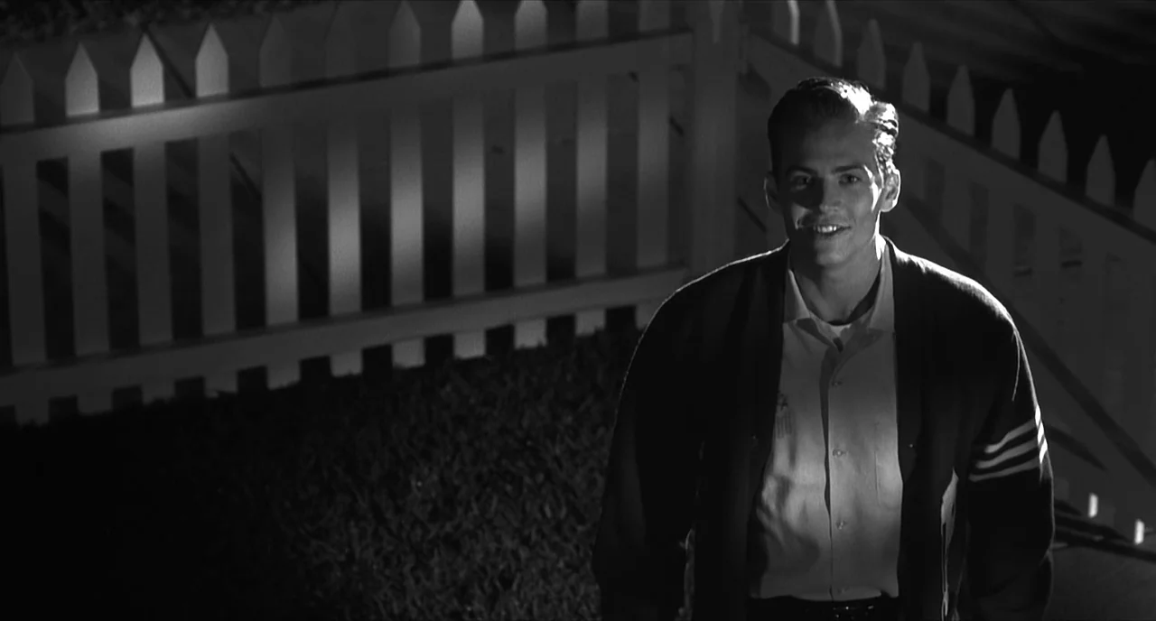 Pleasantville (1998) movie still: medium — A night scene featuring a wooden picket fence backdrop in a suburban setting. A young man stands in front…