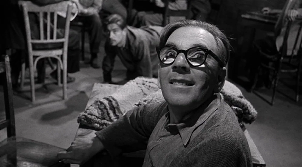 Suddenly, Last Summer (1959) movie still: medium — A dimly lit room with wooden tables and chairs in a crowded, rustic setting. A middle-aged man in…