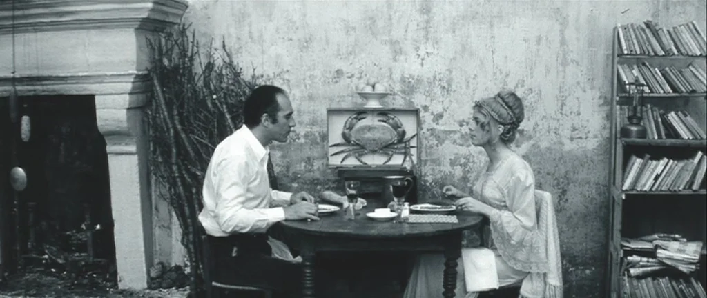 Les Créatures (1966) movie still: fullbody — A sparse interior with a weathered wall, a stone fireplace, and a wooden table. A man and a woman sit…