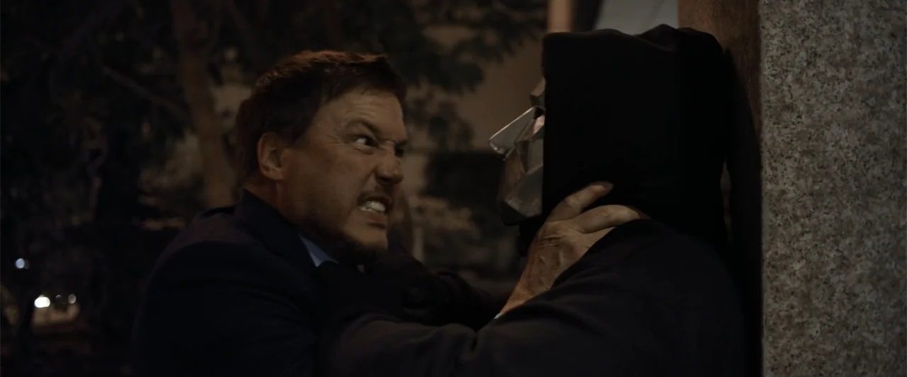 Init!ation (2021) movie still: medium — A dark exterior setting at night featuring a textured stone wall in the background. A man in a suit grasps a…