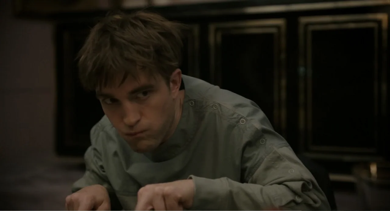 Mickey 17 (2025) movie still: medium — A dimly lit room features a man sitting at a dark surface indoors. The subject focuses his gaze while eating…
