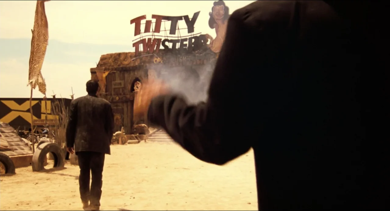 From Dusk Till Dawn (1996) movie still: medium — A dusty desert area sits in front of a wooden structure with a neon sign. A man in a dark suit…