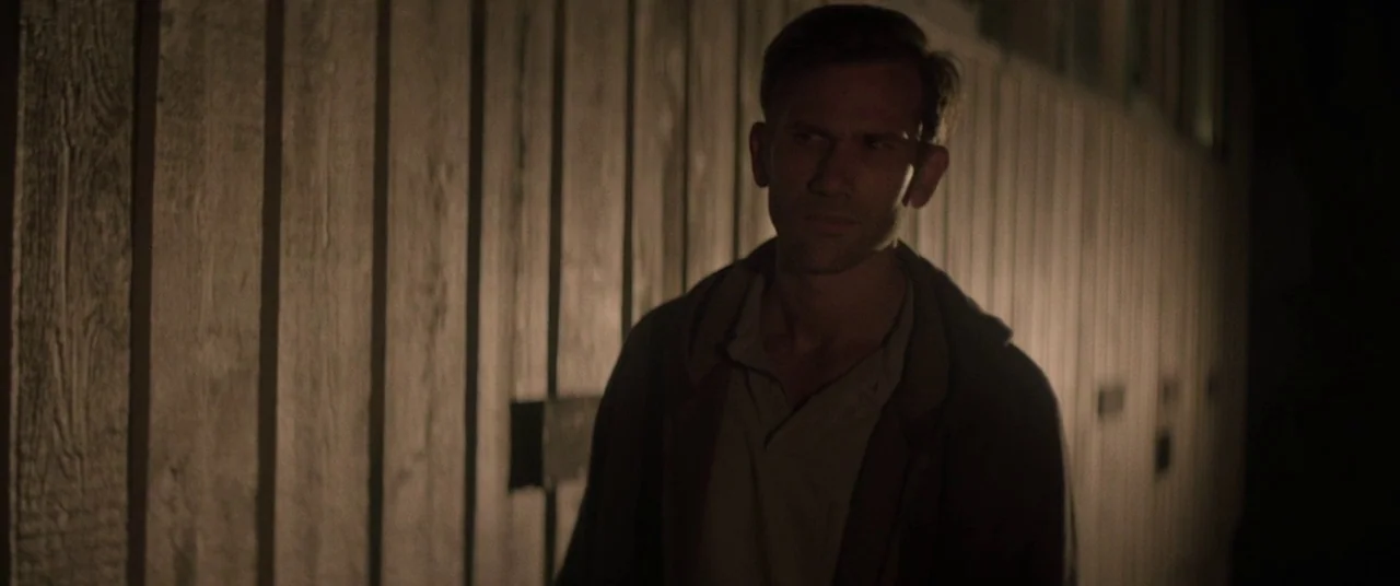 The Endless (2017) movie still: medium — A man walks alongside a dark, vertical wood-planked structure at night. He moves steadily past the textured…