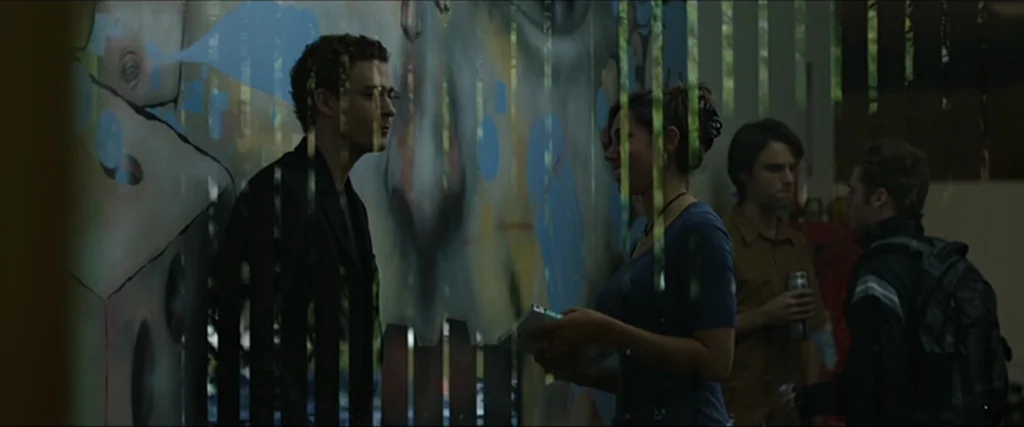 The Social Network movie still: medium — A group of young adults congregates in a building corridor with a mural wall. A man and a woman converse…