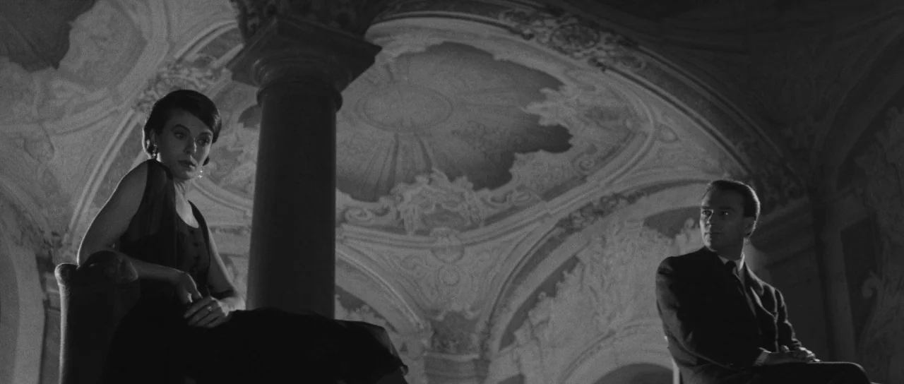 Last Year at Marienbad (1961) movie still: medium — A spacious room features ornate vaulted ceilings with intricate plasterwork and a central…