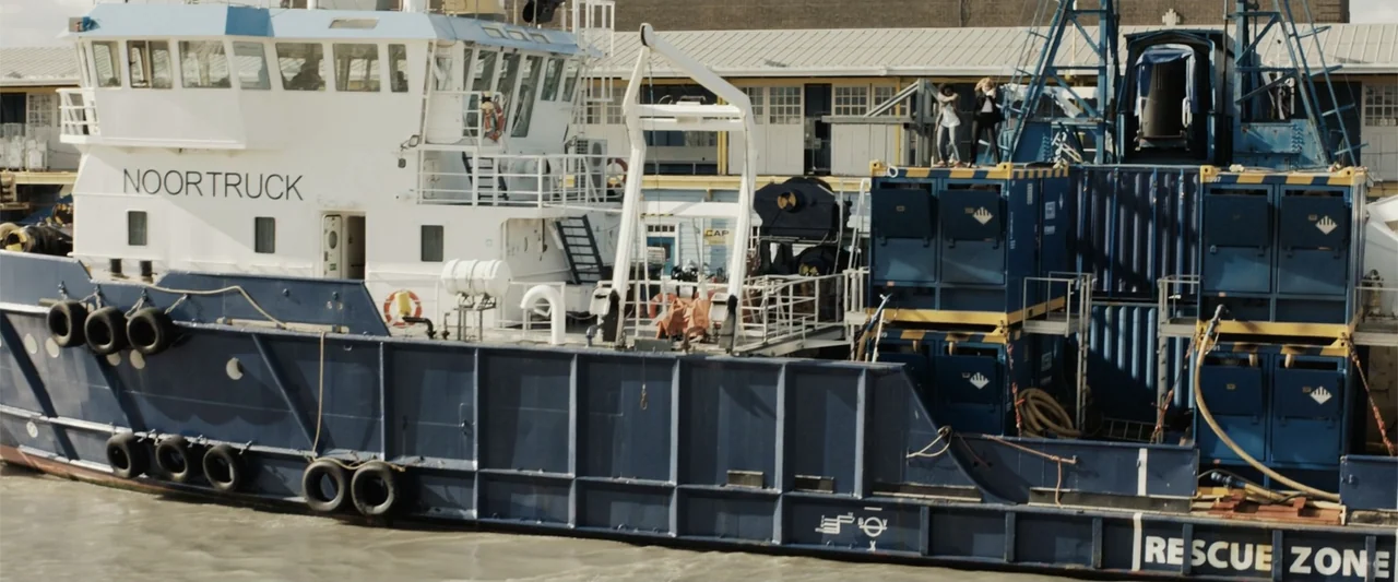 The 355 (2022) movie still: wide — A large industrial supply ship floats on water next to a commercial building complex. Two people stand on the top…