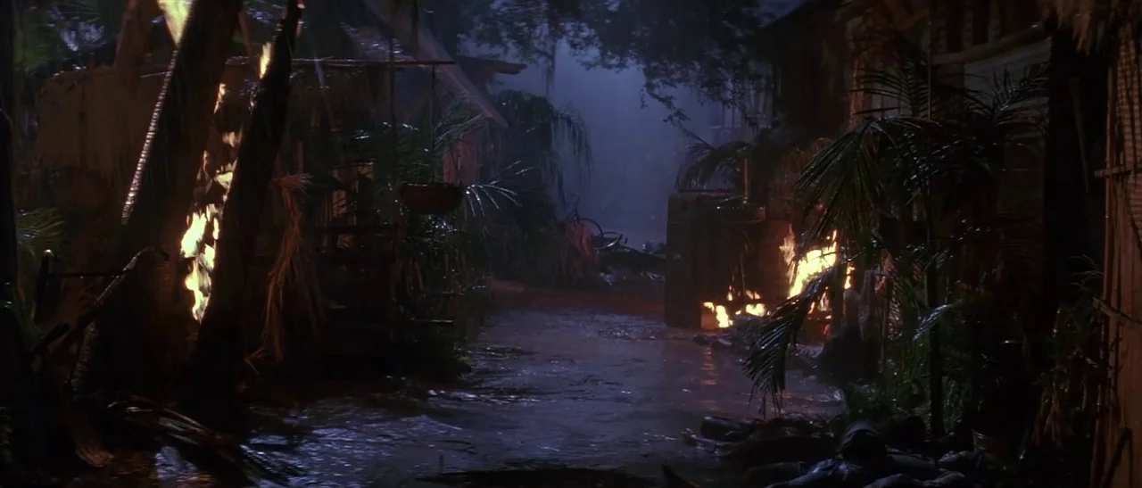 Universal Soldier (1992) movie still: establishing — A dark, rain-swept tropical village at night features wooden buildings engulfed in active…