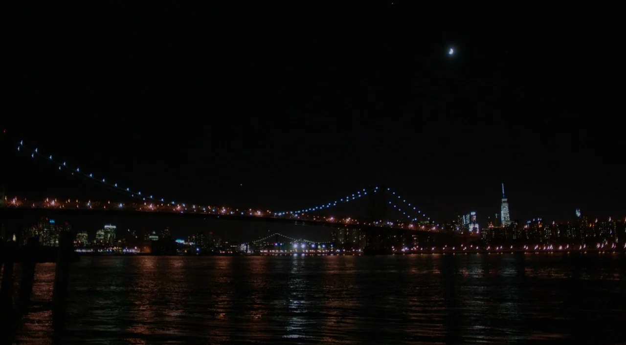 The Big Sick (2017) movie still: establishing — The dark East River flows beneath the illuminated Williamsburg Bridge at night. A crescent moon…