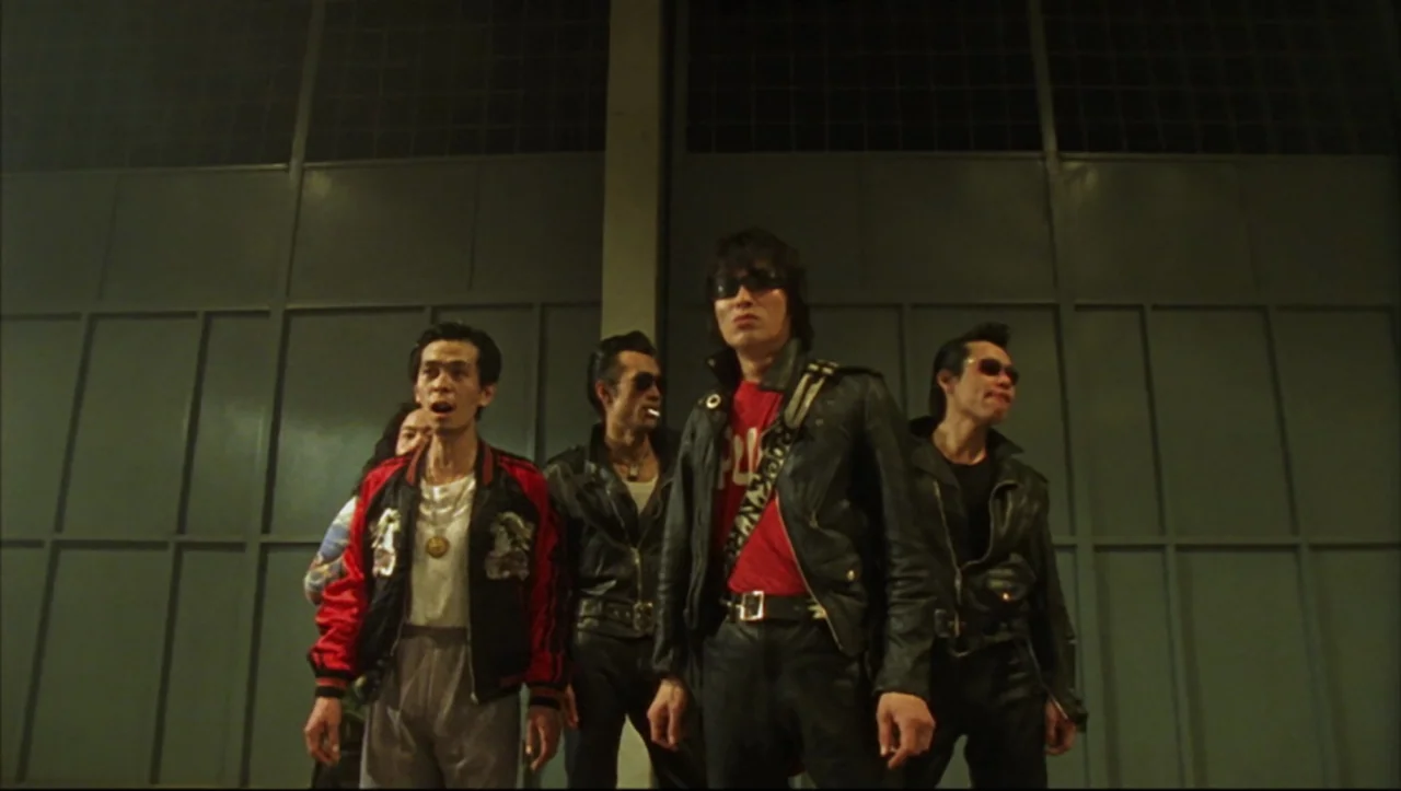 Wild Zero (1999) movie still: fullbody — A group of four men stands before large steel industrial doors at night. The men wear matching leather…