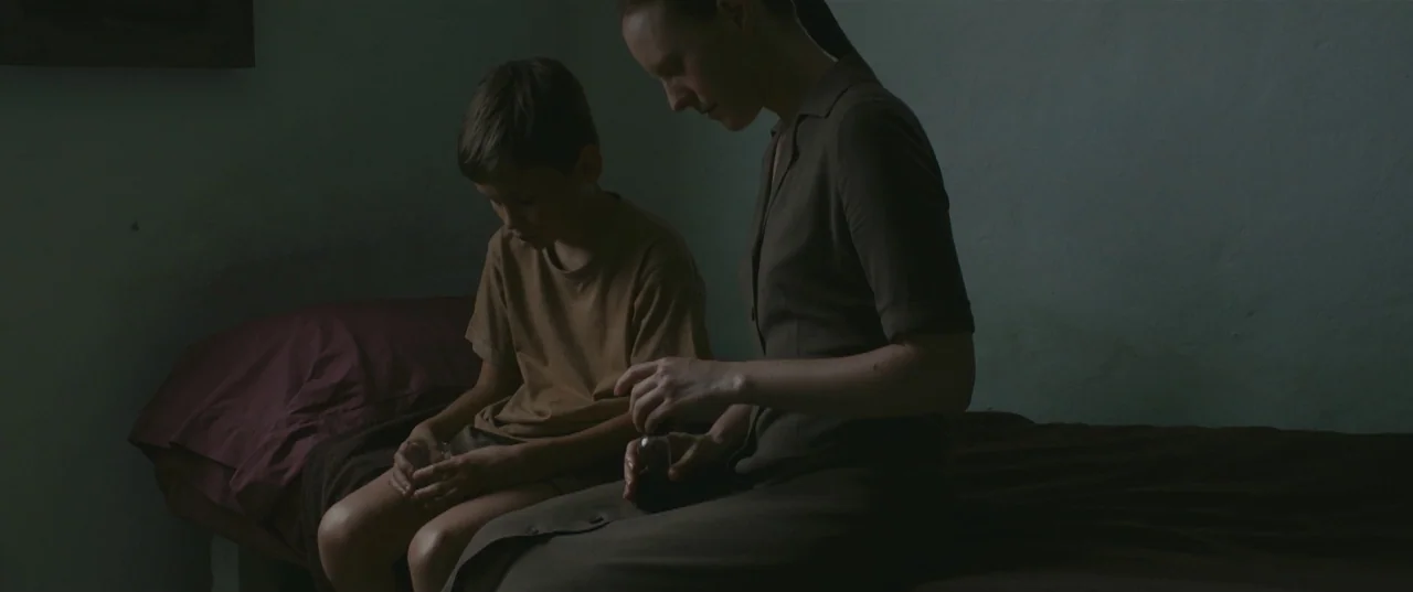 Evolution (2015) movie still: medium — A dimly lit, sparse room with a bed covered in dark linens. A woman sits beside a boy and carefully pours…