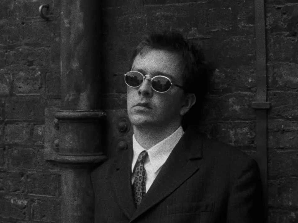 Following (1998) movie still: medium — A man stands against a dark, weathered brick wall with an attached metal pipe. The man wears a suit while…