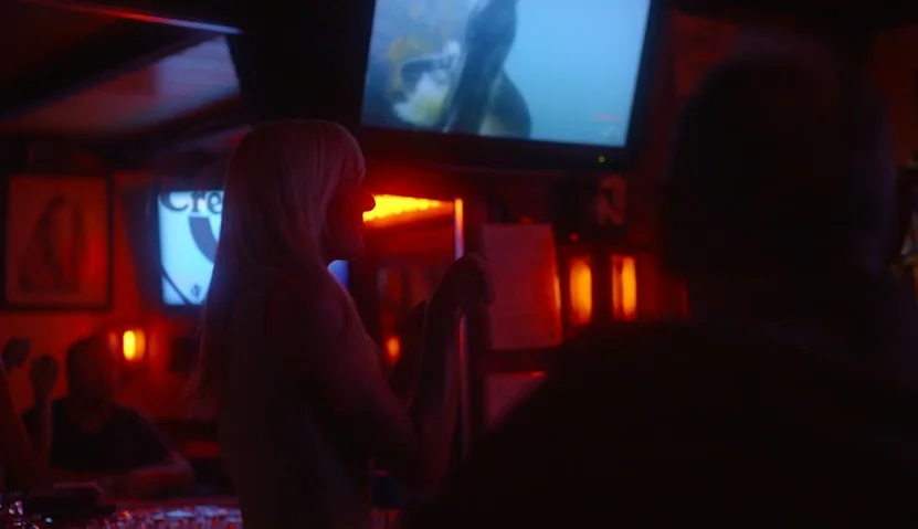 Kate Plays Christine (2016) movie still: medium — A dark bar interior with red neon accent lighting and multiple glowing television screens. A…