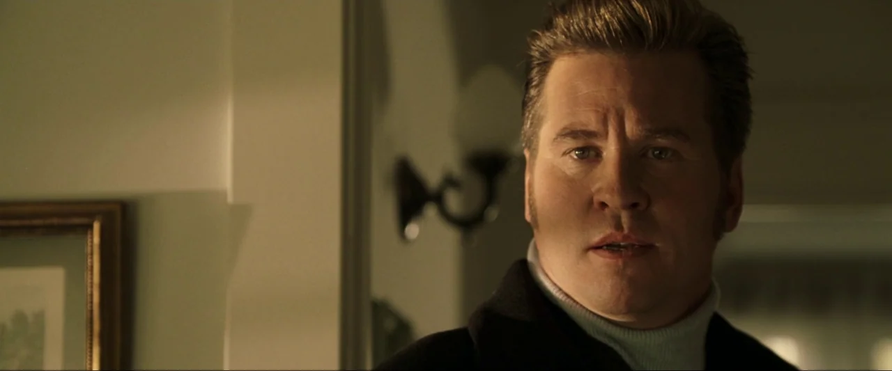 Kiss Kiss Bang Bang (2005) movie still: closeup — A dimly lit indoor hallway with a wall-mounted lamp and a framed picture. A man stands facing…