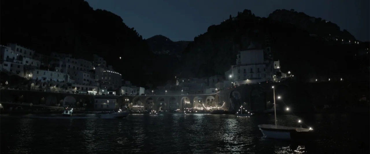 The Equalizer 3 (2023) movie still: establishing — A coastal village rests at the base of dark cliffs at night. Small watercraft move across the…