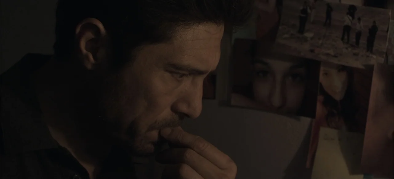 About the Deaths (2016) movie still: closeup — A dimly lit room features a man standing before a wall of photographs. The man gazes at the images…