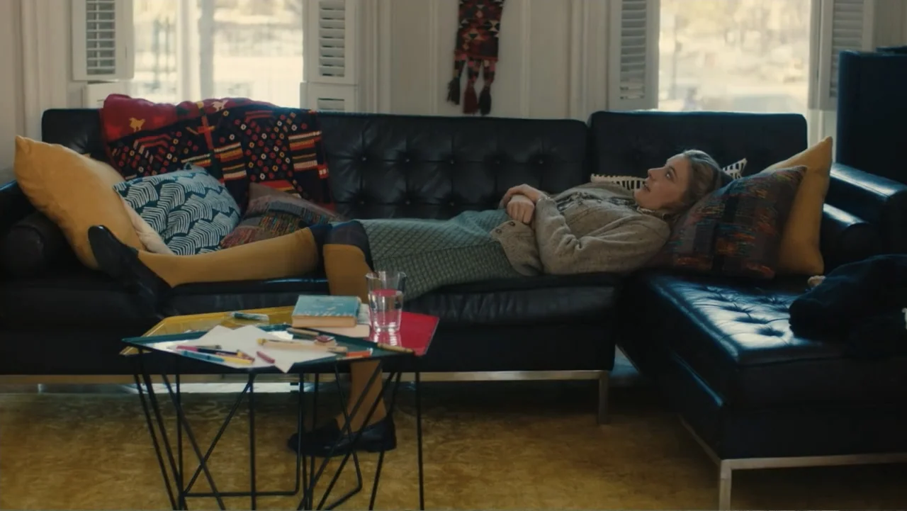 Maggie's Plan (2016) movie still: medium — A living room features a black leather sofa with patterned pillows and mustard-colored textiles. A blonde…