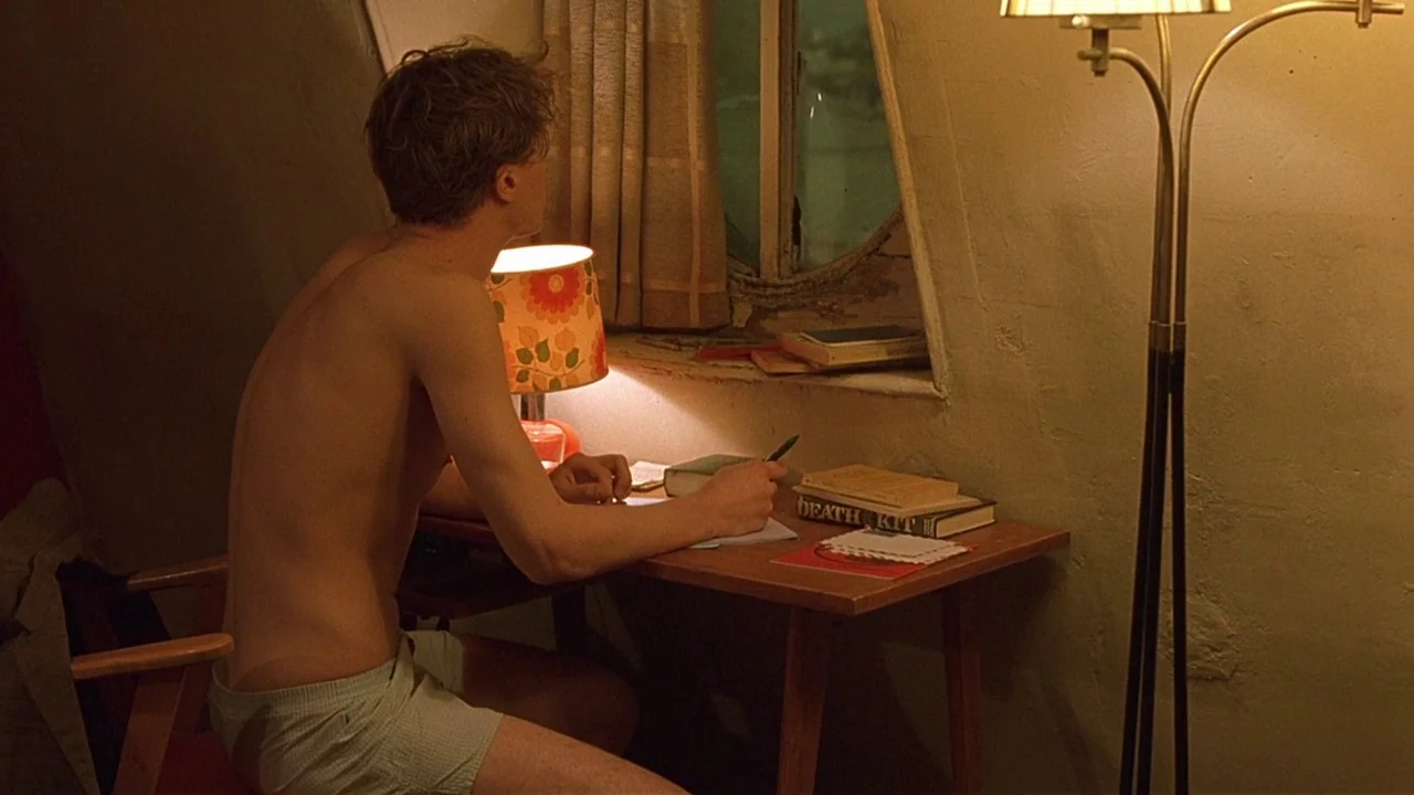 The Dreamers (2003) movie still: medium — An attic room contains a wooden desk placed beneath a small, slanted window. A young man sits at the desk,…
