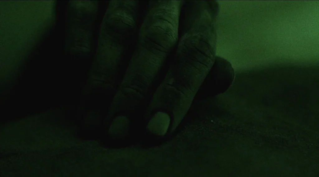 Buried (2010) movie still: closeup — A dark, textured surface of sand is lit with a green monochromatic night vision. A single human hand rests flat…