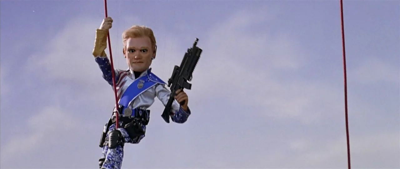 Team America: World Police movie still: fullbody — A marionette puppet dressed in patriotic gear hangs in front of a blue sky. The puppet holds a…