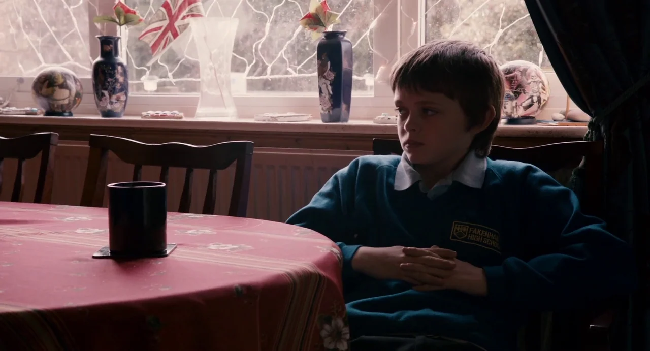 Everyday (2012) movie still: medium — A dining room with a window featuring decorative vases and a flag. A young boy in a school uniform sits at a…