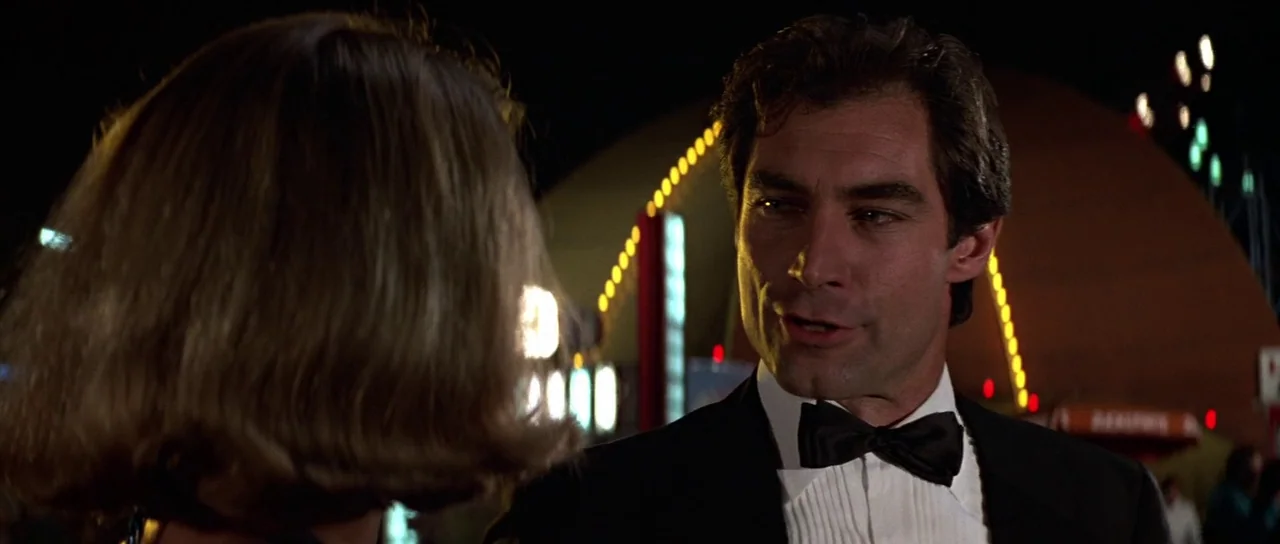 The Living Daylights (1987) movie still: medium — An amusement park at night features bright blurred neon lights in the dark background. A man in…