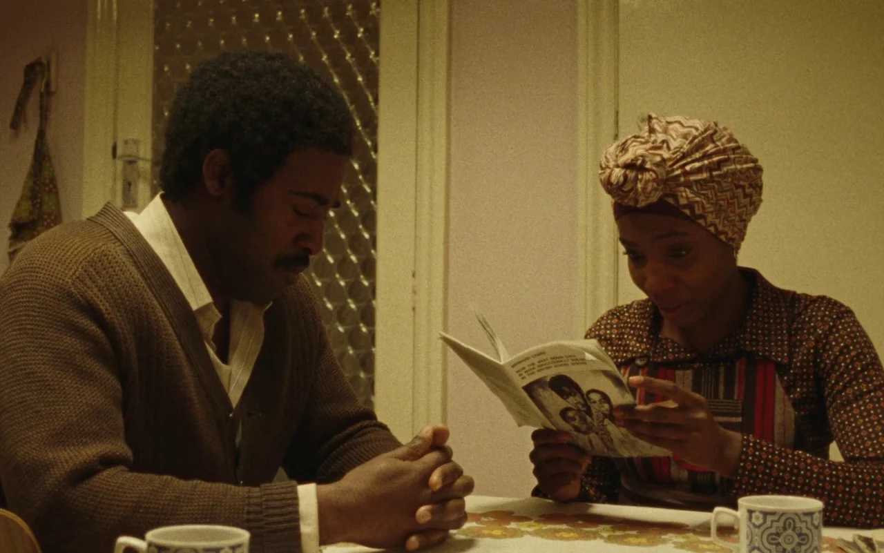 Education (2020) movie still: medium — A man and woman sit at a patterned table inside a warmly lit room. The woman examines a printed pamphlet…