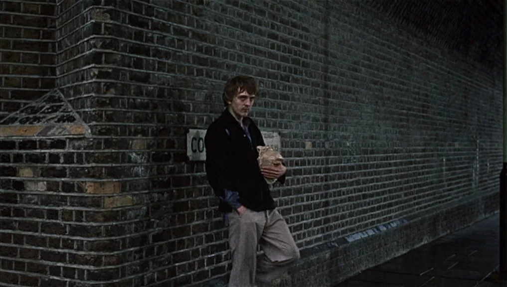 Blow-Up (1966) movie still: fullbody — A dark brick underpass features a weathered wall with an illegible metal street sign. A young man with short…