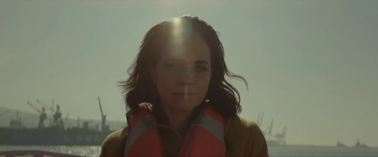 Ema (2019) movie still: medium — A woman stands on a boat with a busy industrial shipyard in the distance. She is wearing a bright orange life vest…