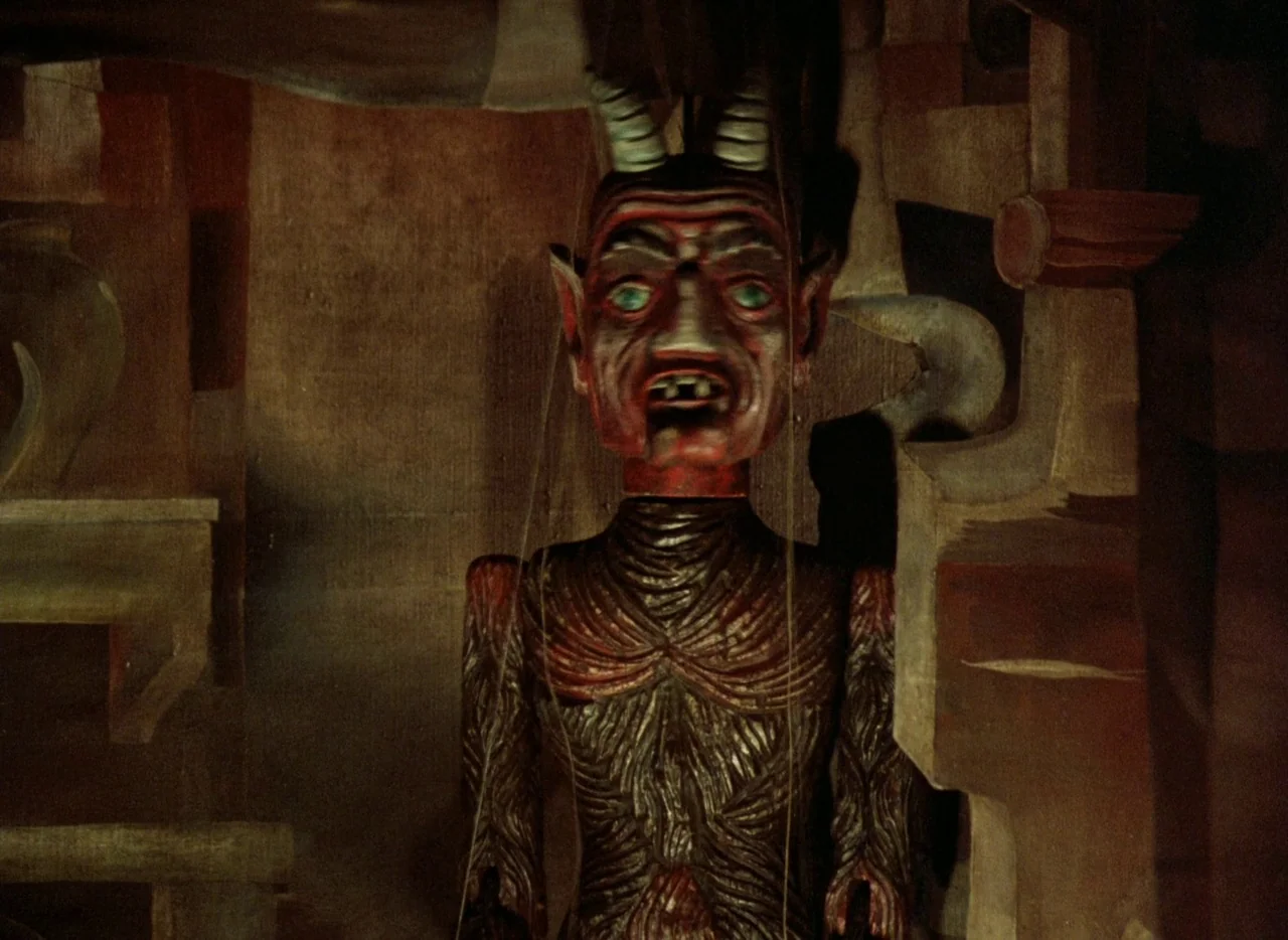 Faust (1994) movie still: medium — A carved wooden devil marionette hangs inside a dark and dimly lit puppet theater. Thin control wires extend…