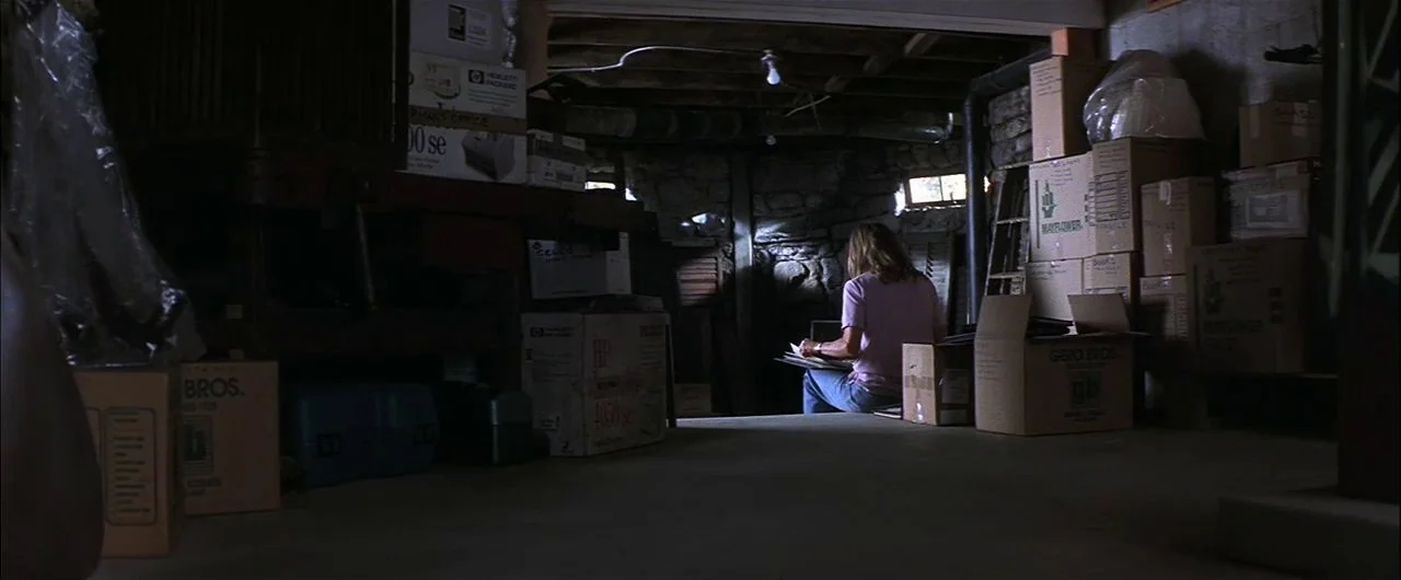 What Lies Beneath (2000) movie still: wide — A dimly lit basement is filled with stacked cardboard boxes and storage items. A woman sits on the…