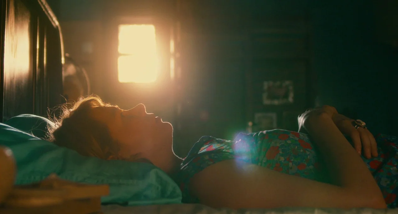 Take This Waltz (2011) movie still: medium — A woman rests on a bed inside a dimly lit bedroom setting. Bright light spills from a window behind…