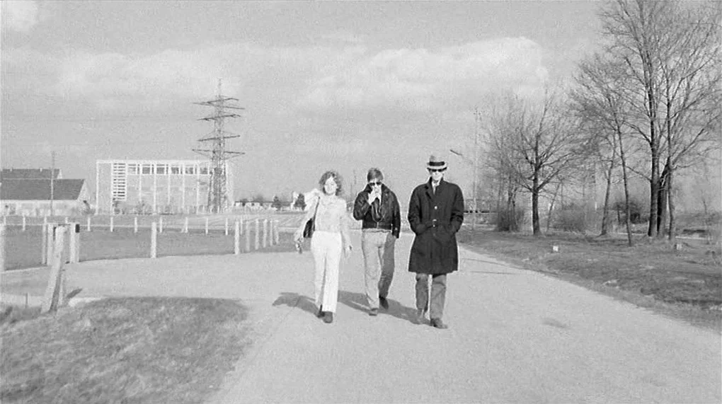 Love Is Colder Than Death (1969) movie still: fullbody — A paved path runs through a sparse landscape with bare trees and distant buildings. Three…